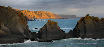 Cornish cliffs This landscape photograph captures the Cornish cliffs along the coast of Cornwall in the United Kingdom, with dramatic rock formations rising from the sea. The image was taken during the evening in autumn, as suggested by the warm, low-angle sunlight illuminating the rugged cliff faces and the rich golden hues spreading across the rural landscape. The natural coastline, shaped by years of erosion, forms distinctive triangular shapes in the foreground, and the calm sea contrasts with the imposing cliffs behind. This scene exemplifies the unspoiled nature of the area, highlighting the wild beauty of rural Cornwall by the sea.